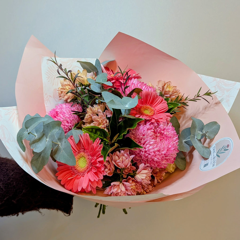 A vibrant bouquet of pink and red gerbera daisies with green eucalyptus leaves, wrapped in light pink paper with a small round floral sticker on the right side.