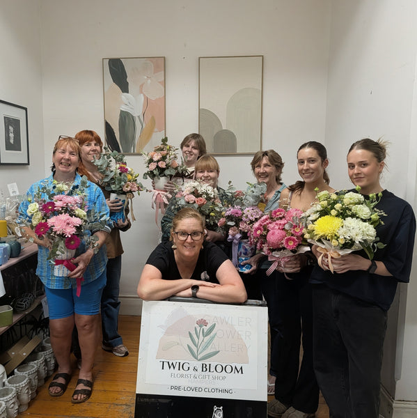 A group of women standing indoors holding bouquets of flowers, with one woman in the front center leaning on a sign that reads 'TWIG & BLOOM FLORIST & GIFT SHOP' and 'PRE LOVED CLOTHING.' This is a real indoor environment with people present, so the correct subject_type is Lifestyle rather than Product/Packaging.