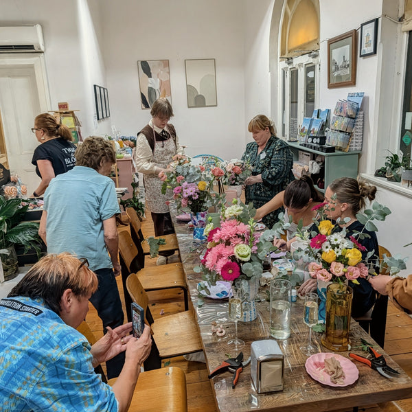 A group of people gathered around a long table in a bright room, arranging colorful floral centerpieces. The scene shows multiple individuals working together in a real indoor environment, so the subject type is classified as Lifestyle.