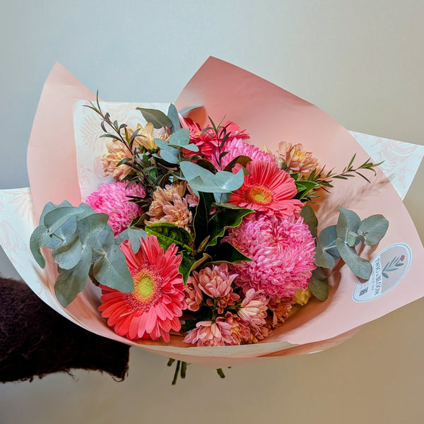 A vibrant bouquet of pink and red gerbera daisies with green eucalyptus leaves, wrapped in light pink paper with a small round floral sticker on the right side.