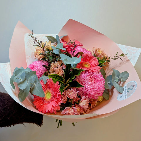 A vibrant bouquet of pink and red gerbera daisies with green eucalyptus leaves, wrapped in light pink paper with a small round floral sticker on the right side.
