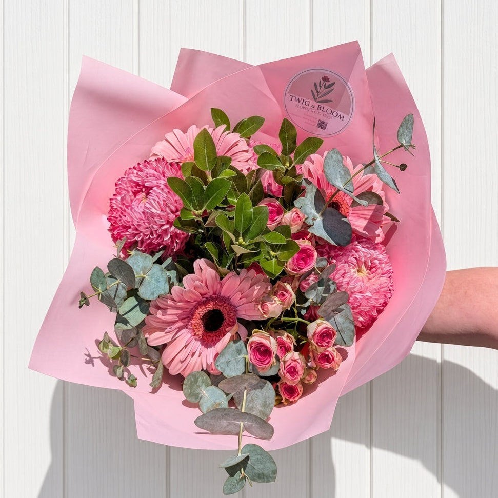 A hand holding a fresh flower bouquet wrapped in pink paper, featuring pink gerbera daisies, pink chrysanthemums, and green foliage, set against a white vertical wood-paneled wall.