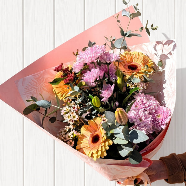 A vibrant mixed flower bouquet wrapped in pink patterned paper, held by a hand against a white vertical wood paneled wall. The bouquet features yellow and pink gerbera daisies, pink chrysanthemums, eucalyptus sprigs, and small white filler flowers.
