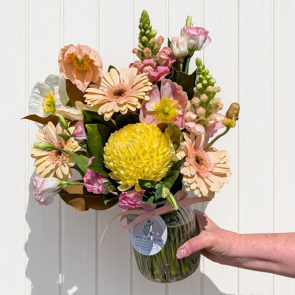 A hand holding a vibrant mixed flower bouquet in a ribbed glass jar vase against a white paneled wall. The bouquet features a large yellow chrysanthemum, peach and pink gerbera daisies, pink snapdragons, and green foliage.