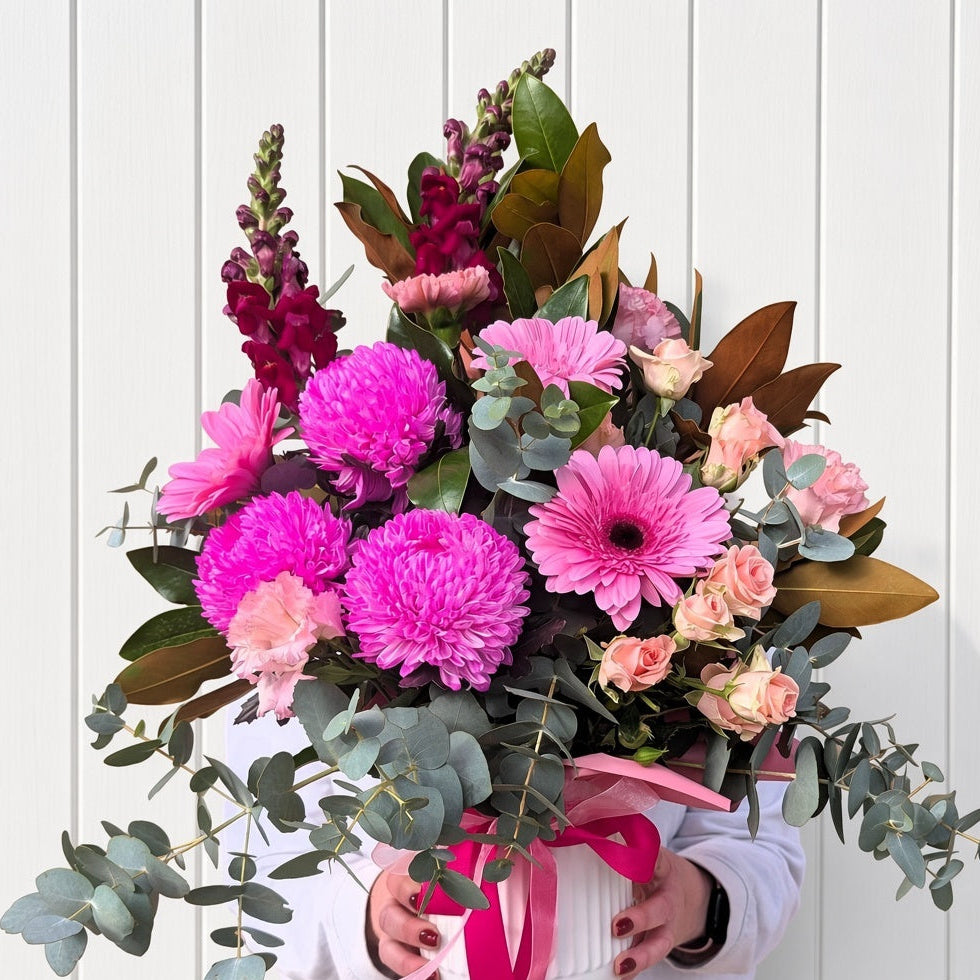 A vibrant floral arrangement held by a person wearing a white top, featuring bright pink chrysanthemums, pink gerbera daisies, pink roses, burgundy snapdragons, and green eucalyptus leaves, set against a white vertical paneled wall.