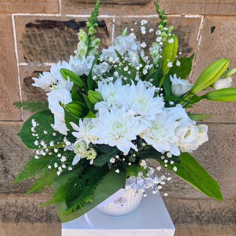 Bouquet of white flowers with green leaves on a white pedestal against a brick wall.