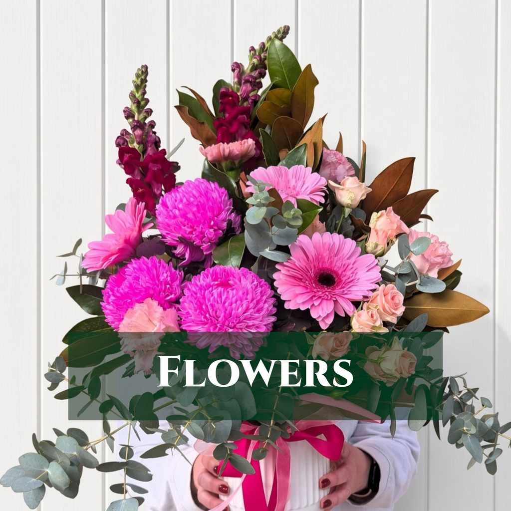 A vibrant floral arrangement featuring bright pink chrysanthemums, pink gerbera daisies, magenta snapdragons, soft pink roses, and green foliage, held by a person wearing a white top and holding a pink ribbon. The background is a white vertical paneled wall, and a dark green banner with the word 'FLOWERS' is overlaid across the center.