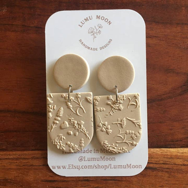 Pair of beige earrings with floral patterns on a LUMU MOON branded card, set against a wooden background.