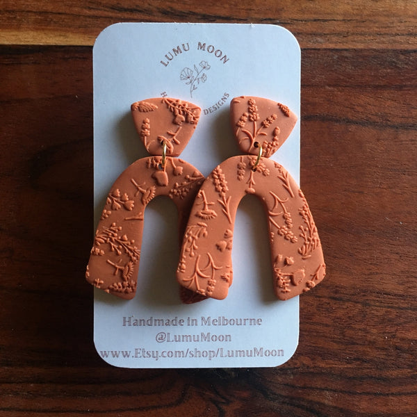 Pair of terracotta-colored earrings on a LUMU MOON branded card against a wooden background.
