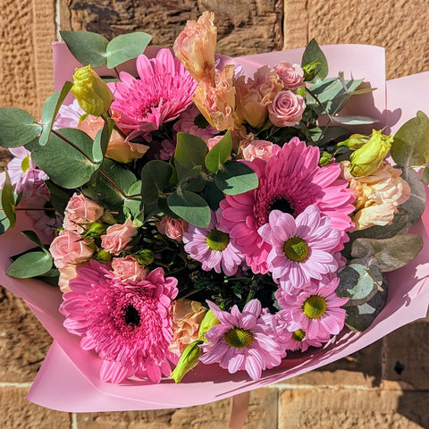 A bouquet of flowers including pink gerberas, chrysanthemums, and spray roses with green foliage, wrapped in pink paper.