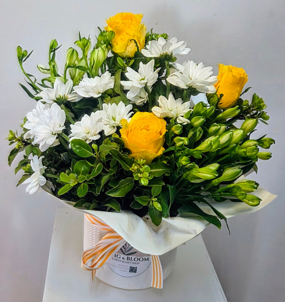 Arrangement of yellow and white flowers with greenery in a white ceramic container, branded 'Twig & Bloom'.