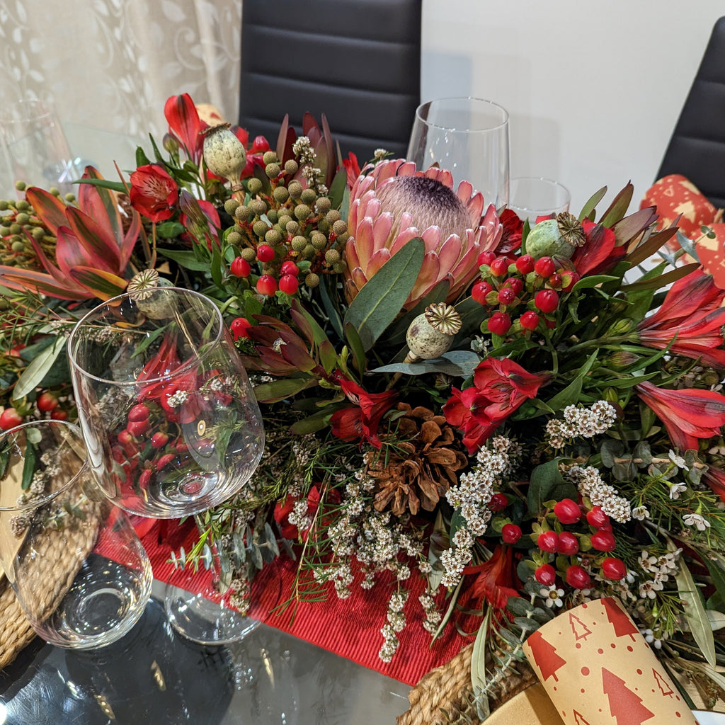 A festive Christmas table arrangement featuring a variety of red and green flowers, foliage, and decorative elements placed on a table with glassware.