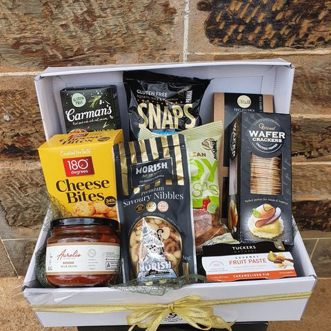 A gift basket containing various snack items and packaged foods, arranged in a white hamper box with a golden ribbon.
